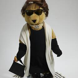 Light brown bear wears black hat, sunglasses, dress, full-length gloves, bag, shoes, white pearls and scarf.