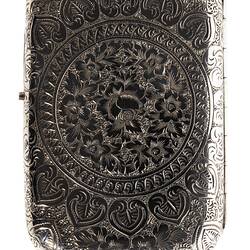Back of heavily engraved silver cigarette case with central text. Rectangular with rounded corners.