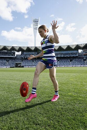 Female footballer wearing Geelong guernsey kicking a football on a football ground. Grandstand behind.