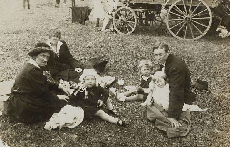 Two women, a man and 3 children picnicking on grass. Wagon wheels visible in background.