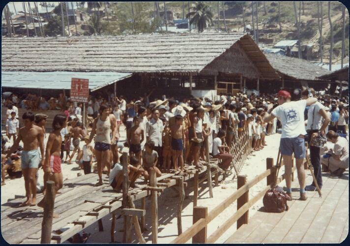 Television crew visiting a Vietnamese refugee camp. Busy scene of many people in and around wooden huts built on sand.