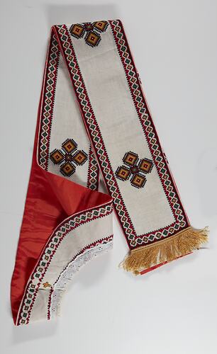 Cream fabric with gold fringe at each end, white lace trim. Red, black, green and yellow border, embroidered geometric star design along length. Red lining on reverse.