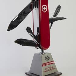 Model of muti-tool on stand. Red handle with all tools out. Large blade, short blade, scissors, bottle opener (with flathead screwdriver), a can opener (with flathead screwdriver), and a reamer.