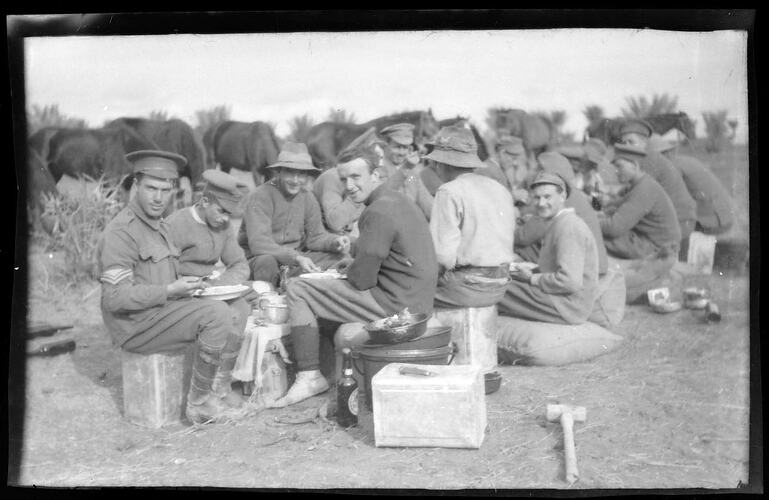 Servicemen seated on boxes and sandbags eating, line of horses behind.