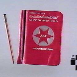 Writing utensil and book with red cover and white text, white circle with red six pointed star in centre.