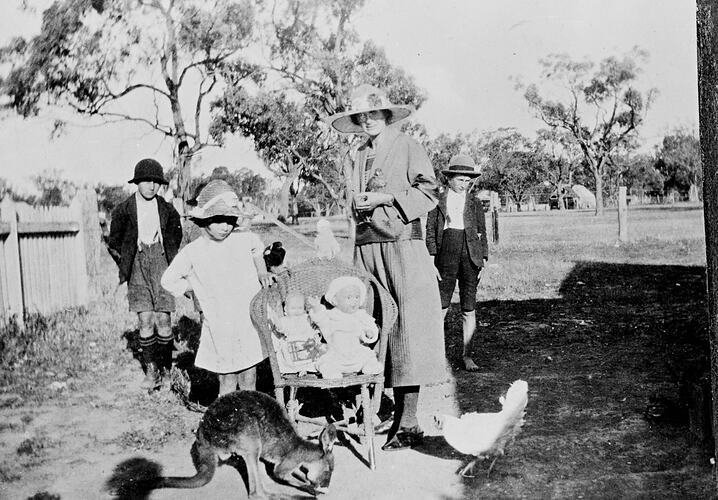 Woman, girl stand either side of chair with two dolls on it. Magpie and galah perch on chair. Two boys in background. Kangaroo and hen in foreground.
