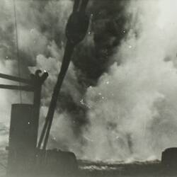 Explosion in water in the middleground, parts of a ship in foreground.