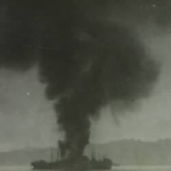 Black plume of smoke rising from ship, land in background.