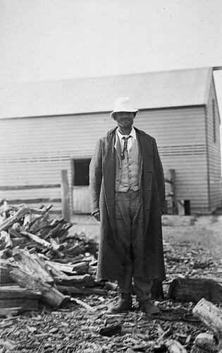 Man stands beside small pile of cut wood. In the background is a large wooden shed.