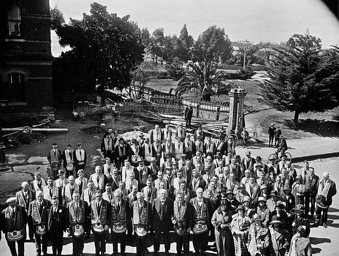 View from height of group. Men in front row where regalia. Trees and gardens behind.