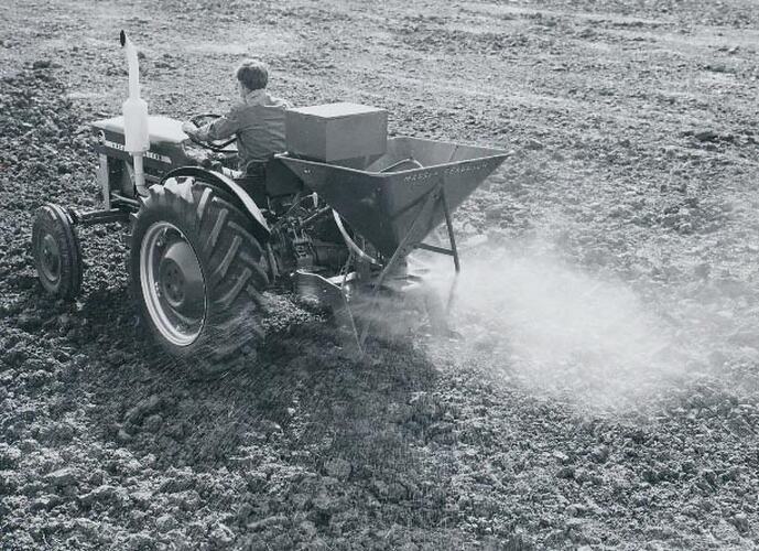 Rear view of a man driving a tractor fitted with a  fertilizer and seed spreader.