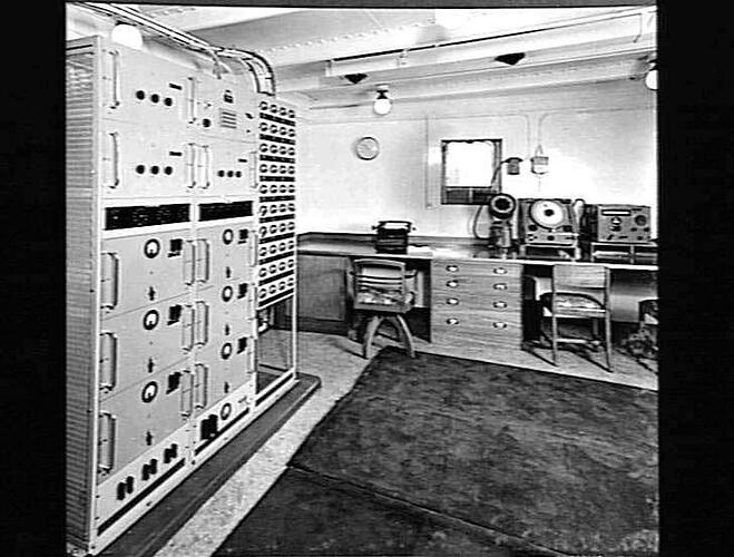 Ship interior. Radio office. Control panel at left. Desk and two chairs against back wall.