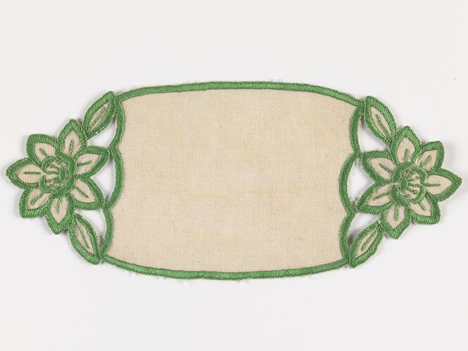 Rectangular cream doily with rounded ends that feature a flower motif with green worked edging.