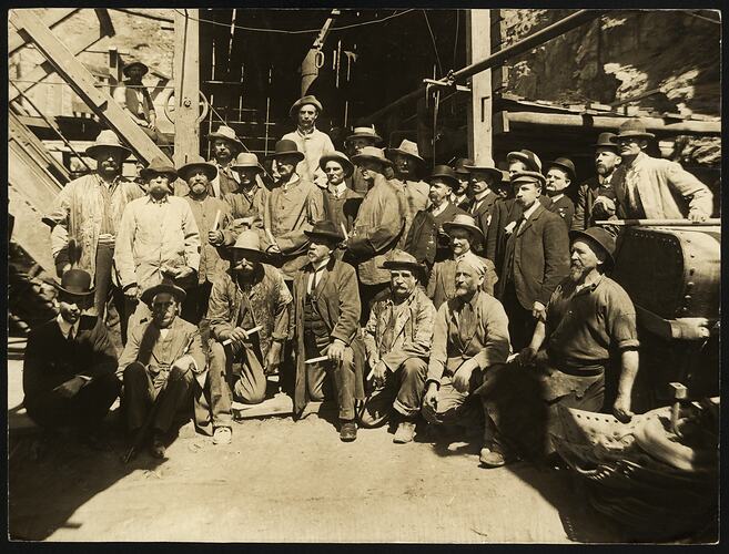 Group of dusty miners at the entrance of a mine. They all wear hats and some hold candles.