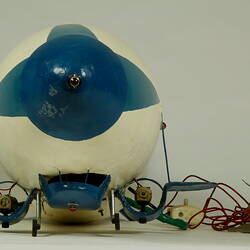 Front view of white model airship, blue nose and wings. Small wheels and cables at base.