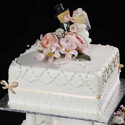 Top tier of white square cake model with pastel ribbons and flowers. Kissing cartoon-style bride and groom.