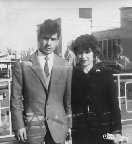 Man and woman pose on ship deck with port behind them.