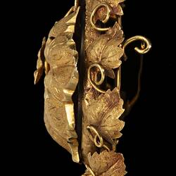 Profile of gold oval brooch. Flying bird across large vine leaf with tendrils. Border of vine leaves, grapes at base.
