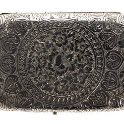Back of heavily engraved silver cigarette case with central text. Rectangular with rounded corners.