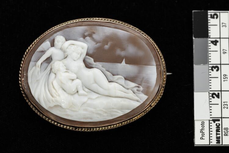 Oval cameo brooch with gold setting. Features reclining figure surrounded by two putto.