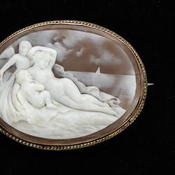 Oval cameo brooch with gold setting. Features reclining figure surrounded by two putto.