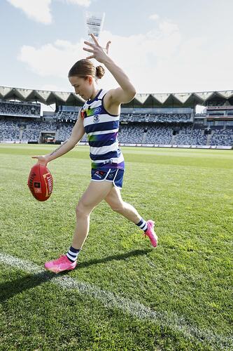 Female footballer wearing Geelong guernsey kicking a football on a football ground. Grandstand behind.