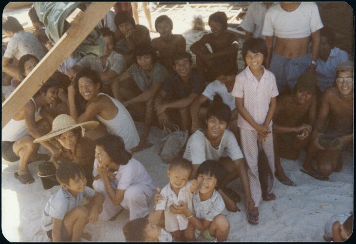 Large group of smiling Vietnamese refugee families, mostly sitting or crouching on sand.