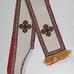 Cream fabric with gold fringe at each end, white lace trim. Red, black, green and yellow border, embroidered geometric star design along length.