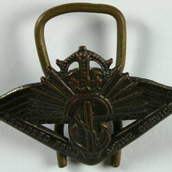 Metal pin with boomerang with embossed text, anchor, rising sun emblem, wings and crown.