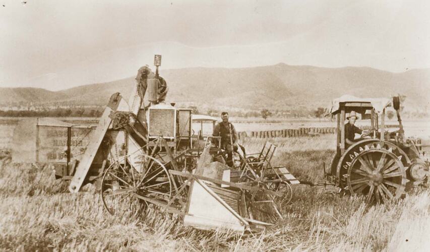 2 men operating a Reaper Thresher, being pulled by a tractor fitted with steel lugged wheels.