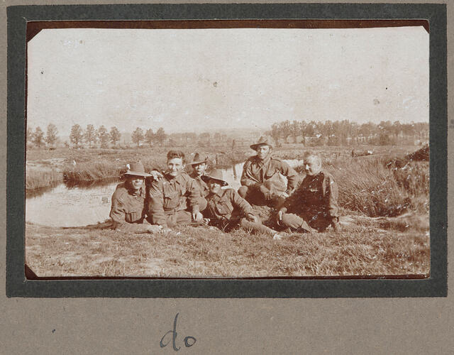 Five servicemen lying on grass in front of a lake, with trees in the background.