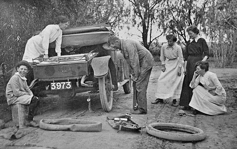 Man pumping up car tyre. Car is jacked up. Tyre and tools lie beside the car. Four women and young boy watch.