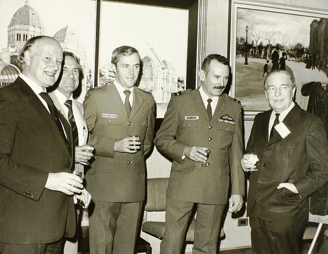 Black and white image of four men in suits, holding drinks in a room with framed paintings on the wall.