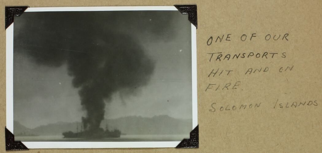 Black plume of smoke rising from ship, land in background.