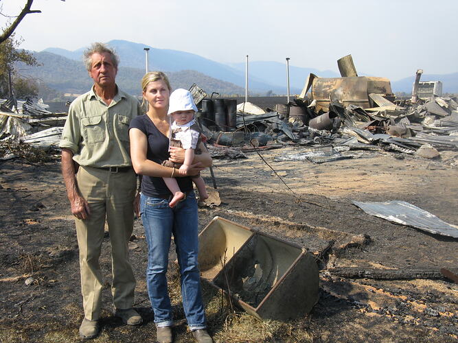 Man, woman and baby stand in front of bushfire damaged collapsed buildings and sheds.