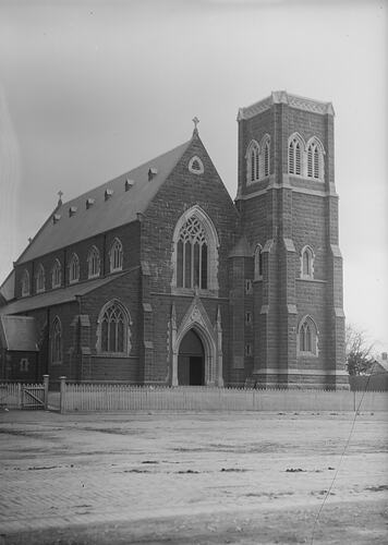 Front view of Roman Catholic church. Entry and belfry tower. Picket fence and dirt road in front.