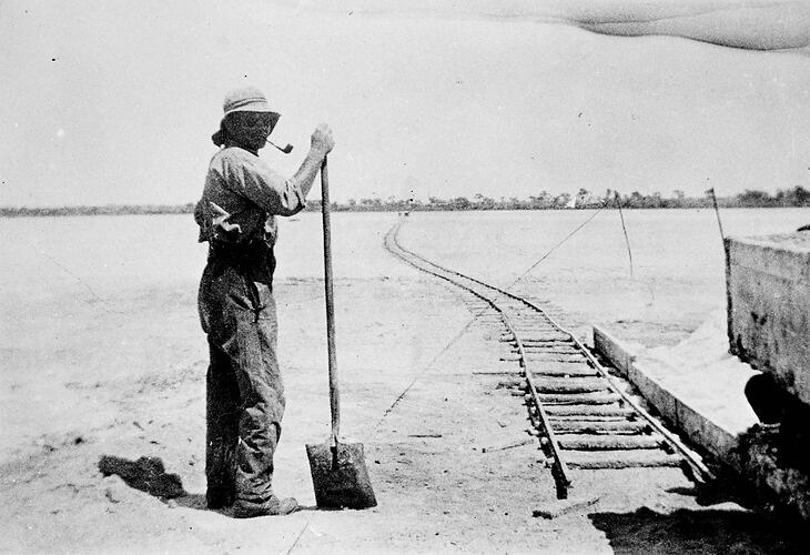 Man standing with shovel beside a long, fairly straight rail line in a flat, treeless setting.