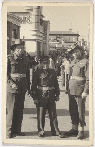 Three men in Australia army uniforms pose with buildings and crowd in the background.