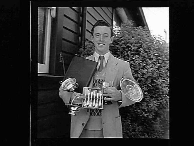 Man wearing a suit holding six trophies and a folder. He stands beside a weatherboard building and bush.