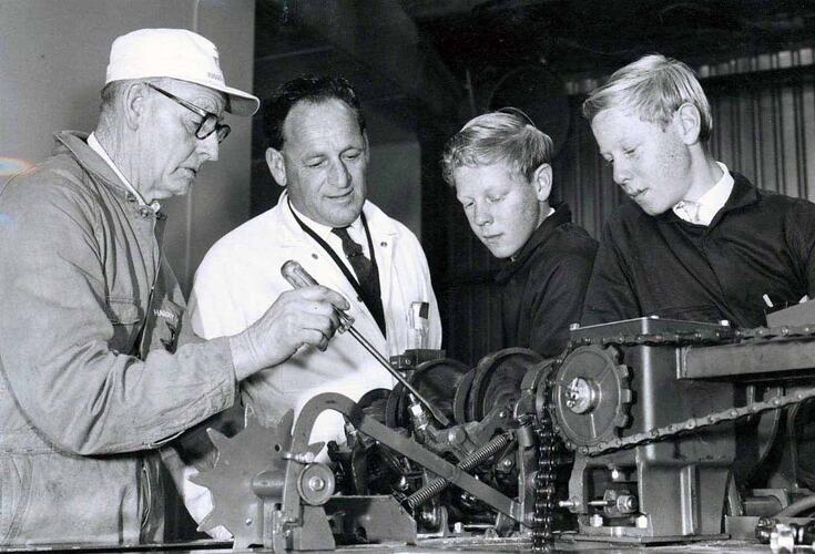 Four men standing behind machine mechanism.