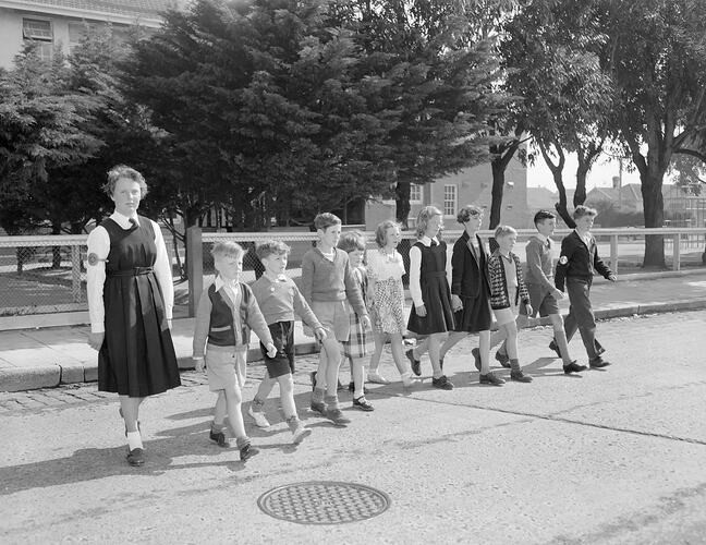 Group of children in a row crossing the road with a woman pictured on the left.