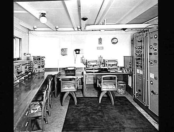 Ship interior. Radio office. Control panel at right. Desk and two chairs at left and along back wall.