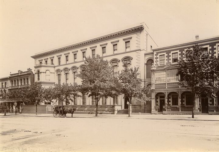 Building exterior from across street. Man stands in doorway. Horsedrawn vehicle outside. Building flanked by trees and a number of terraces style buildings.
