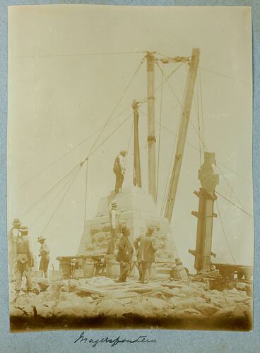 Group of men around a stone monument with rope pullie holding up stone cross.