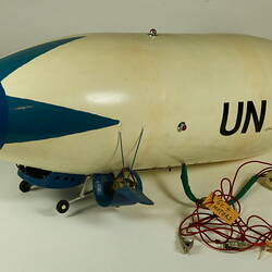 White model airship with blue nose, wings. Black UN on side. Small wheels cables at base. Left profile.