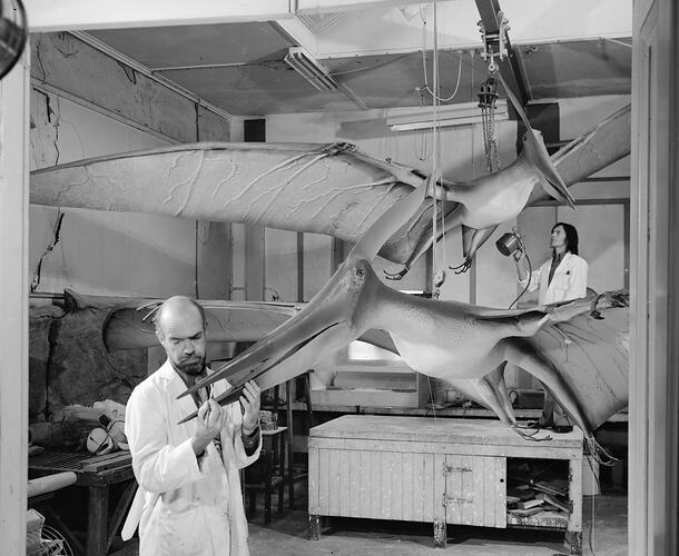 Two men in white coats working on large pterosaur models suspended from workshop ceiling.
