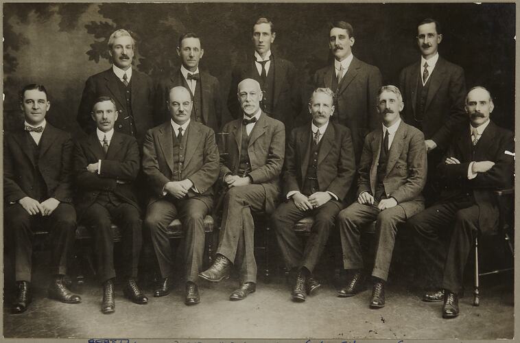 Formal portrait of two rows of men. Back row stand, front row are seated. They wear three piece suits and ties