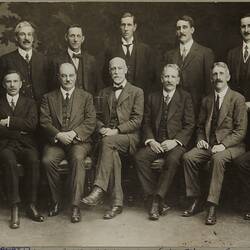 Formal portrait of two rows of men. Back row stand, front row are seated. They wear three piece suits and ties