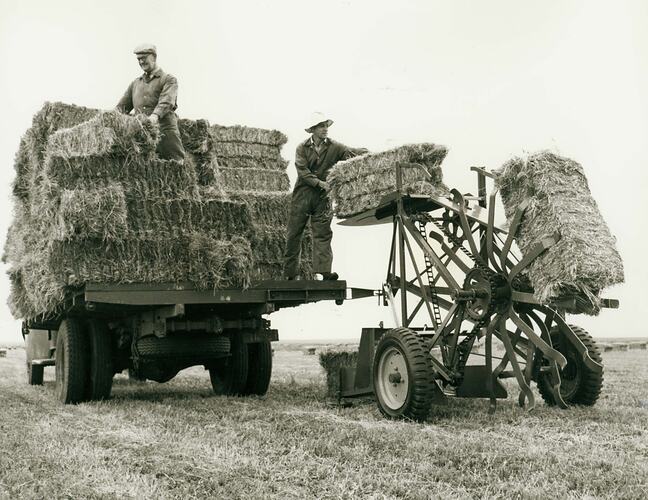 A man lifting a bale of hay from a Sunlifter onto the back of a tray truck loaded with hay bales..