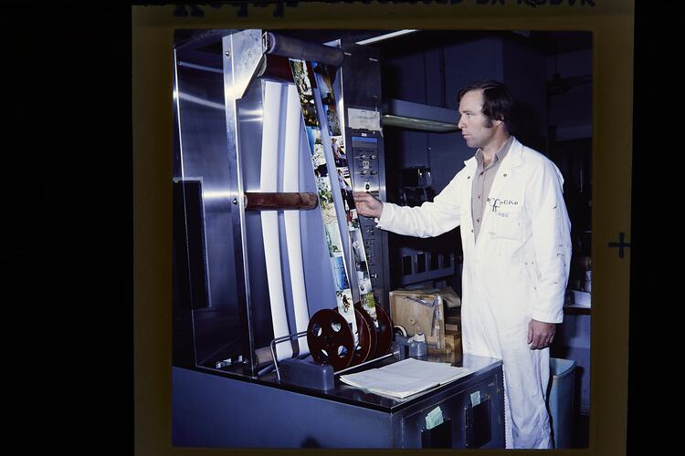 Man in white boiler suit looks at two reels of colour photographs.
